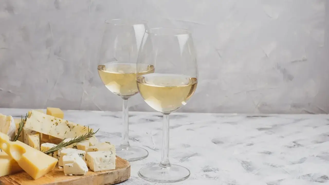 Due calici di Sauvignon Gris dorato accompagnano un tagliere di formaggi misti, tra cui erborinato e stagionato, su un tavolo grigio.