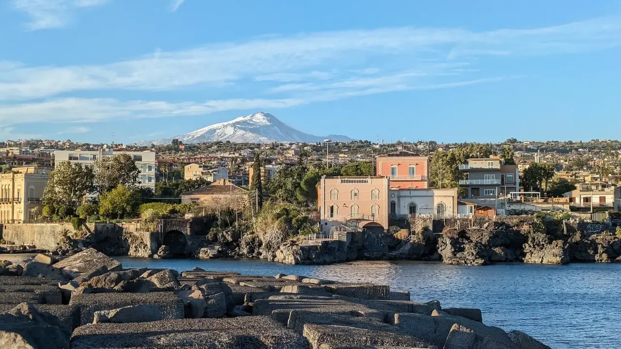 Vista sul mare con blocchi di cemento in primo piano e un panorama di case con l'Etna innevato sullo sfondo, perfetto per un itinerario a Catania.