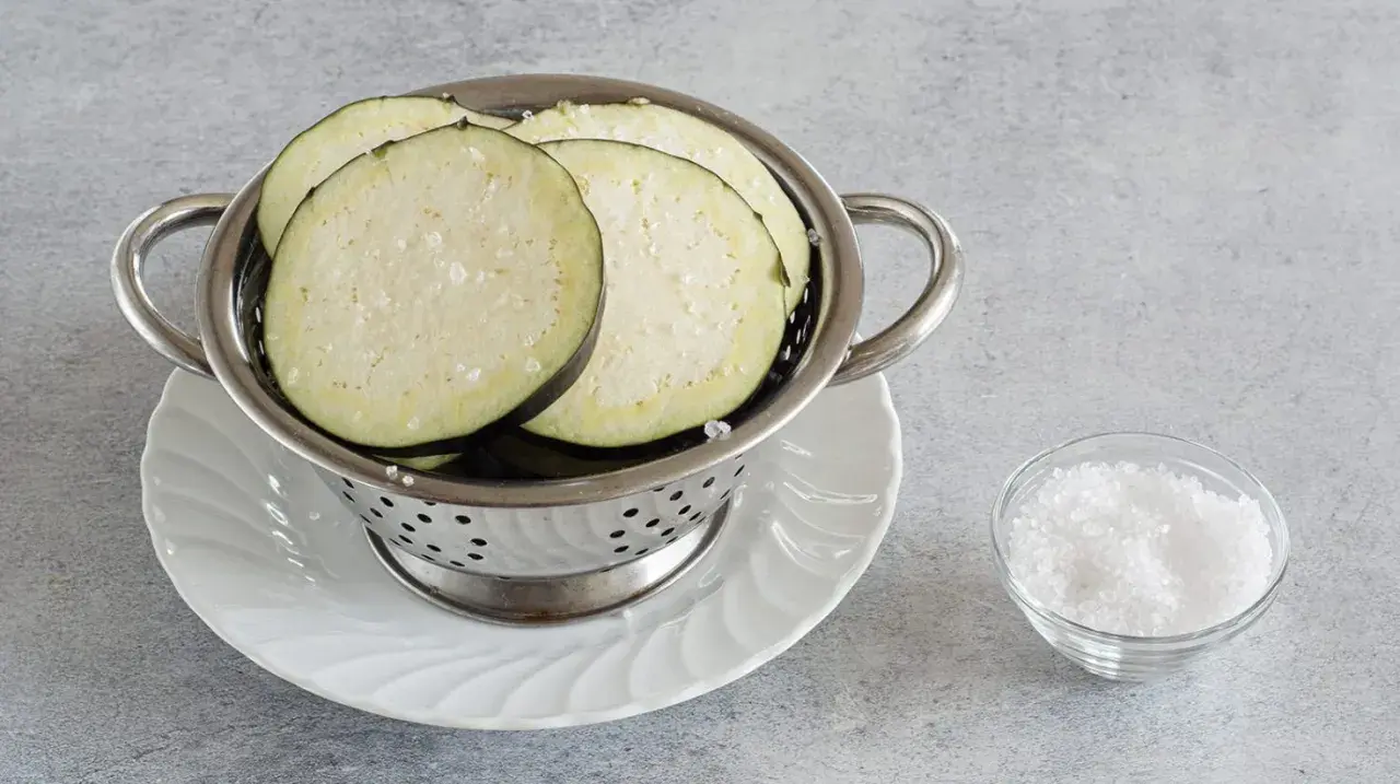 Fette di melanzane salate in un colino, pronte per spurgare. Accanto, una ciotolina di sale grosso.