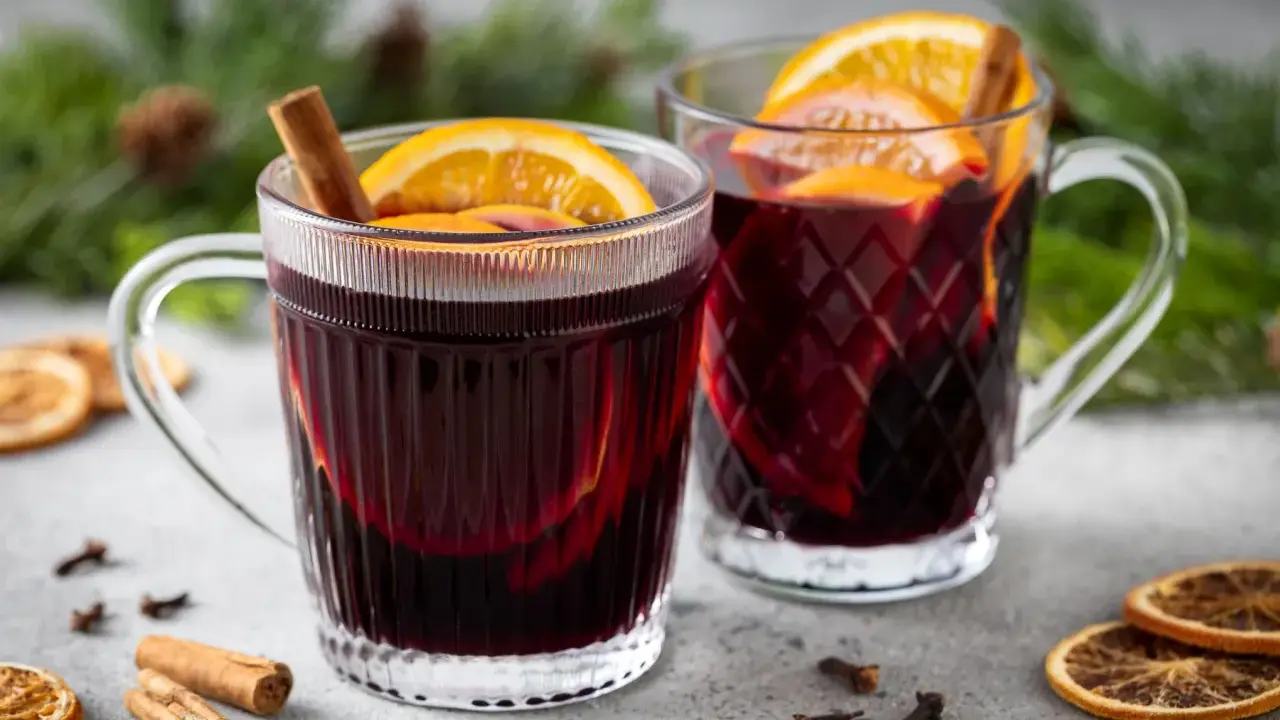 Due tazze fumanti di vin brulé, decorate con arancia e cannella, su un tavolo con spezie e rami di pino.