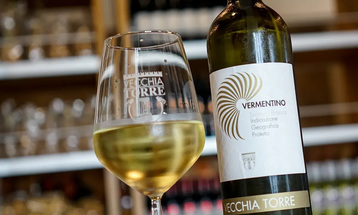 Un bicchiere di vino bianco Vermentino, frizzante e dorato, accanto alla sua bottiglia Vecchia Torre.
