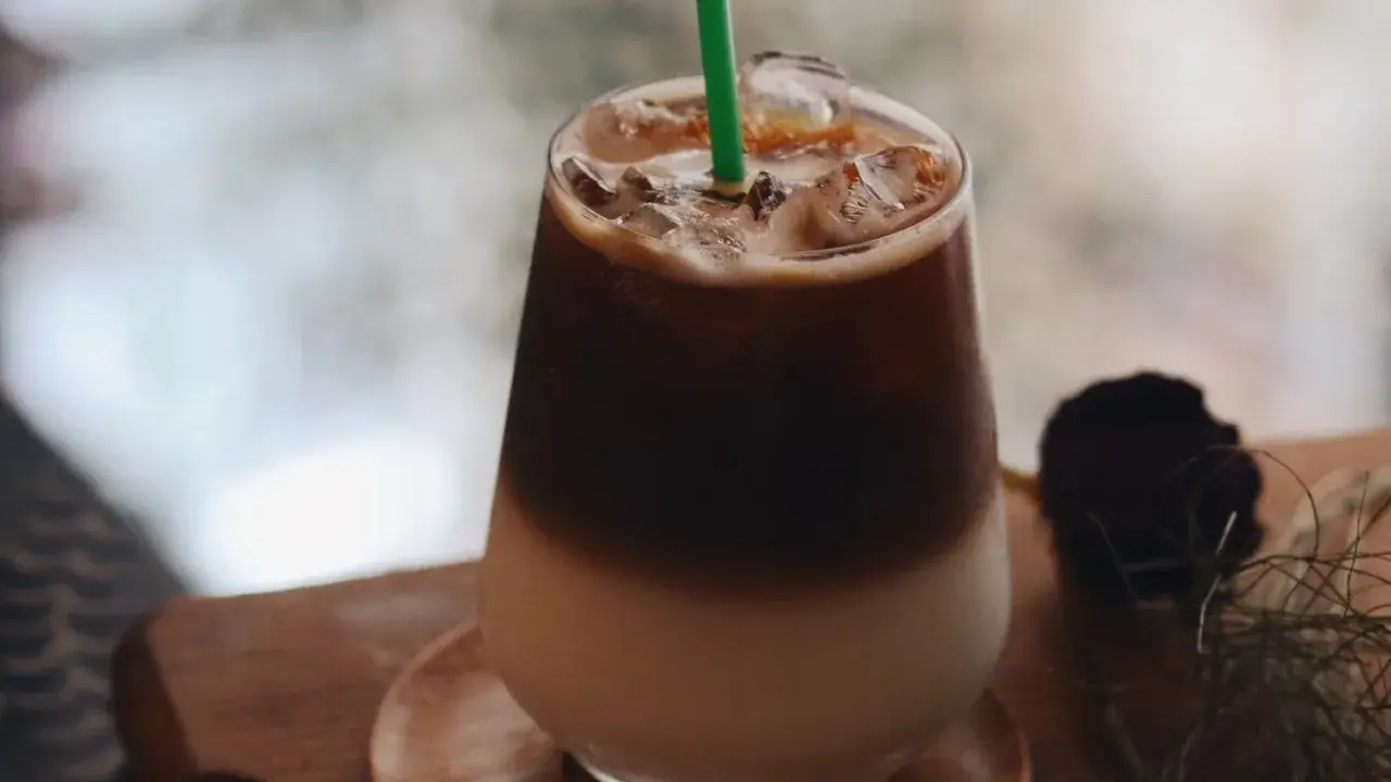 Un bicchiere di **caffè freddo** stratificato con ghiaccio e una cannuccia verde, su un tavolino di legno.