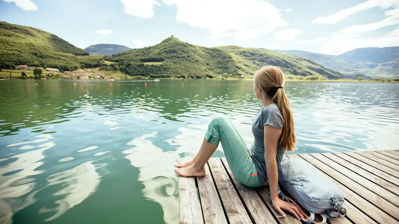 Ragazza con zaino seduta su un pontile di legno, ammira il **lago di Caldaro** e le montagne verdi sotto un cielo azzurro con nuvole bianche.