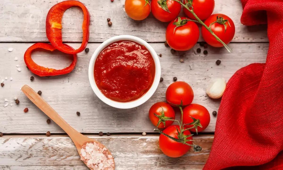 Un ciotola di ketchup fatto in casa, pomodori freschi, peperone rosso, aglio e sale rosa su un tavolo di legno.