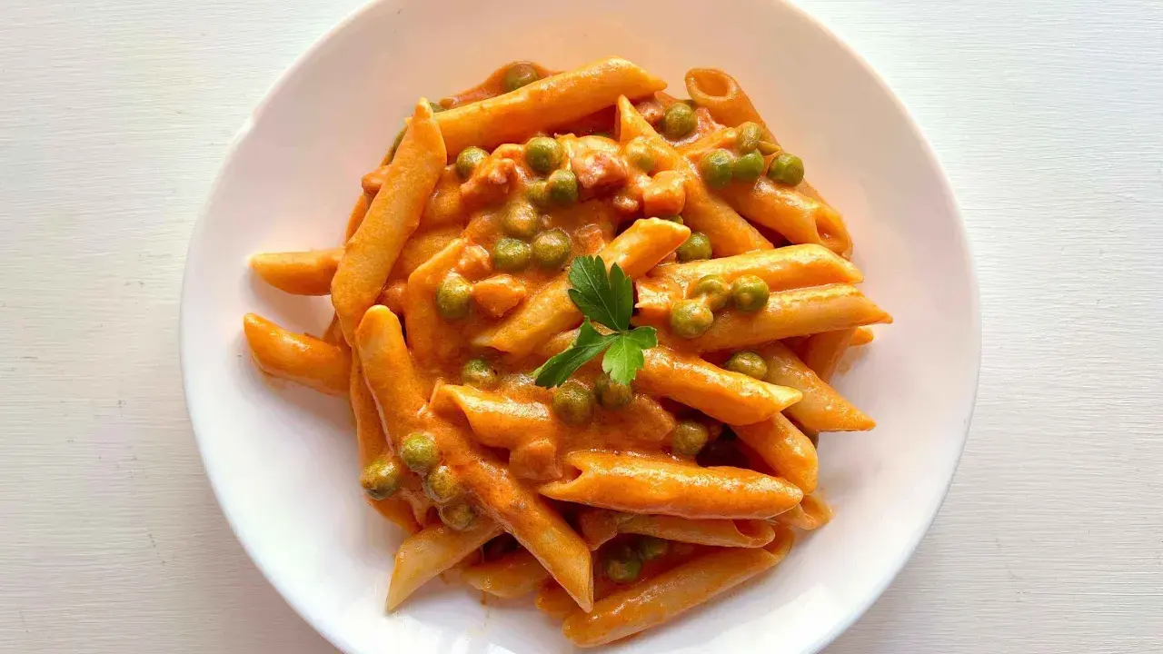 Penne ai 7 peccati: un piatto di pasta cremosa con piselli e un tocco di pomodoro, guarnito con prezzemolo fresco.
