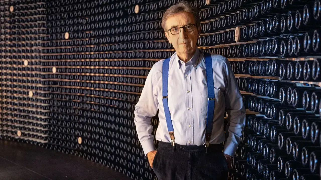 L'enologo Cotarella, con le sue bretelle blu, posa con sicurezza davanti a una parete di bottiglie di vino.