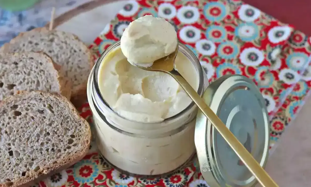 Un cucchiaio dorato preleva una generosa porzione di maionese con uovo sodo da un barattolo di vetro. Fette di pane rustico completano la scena.