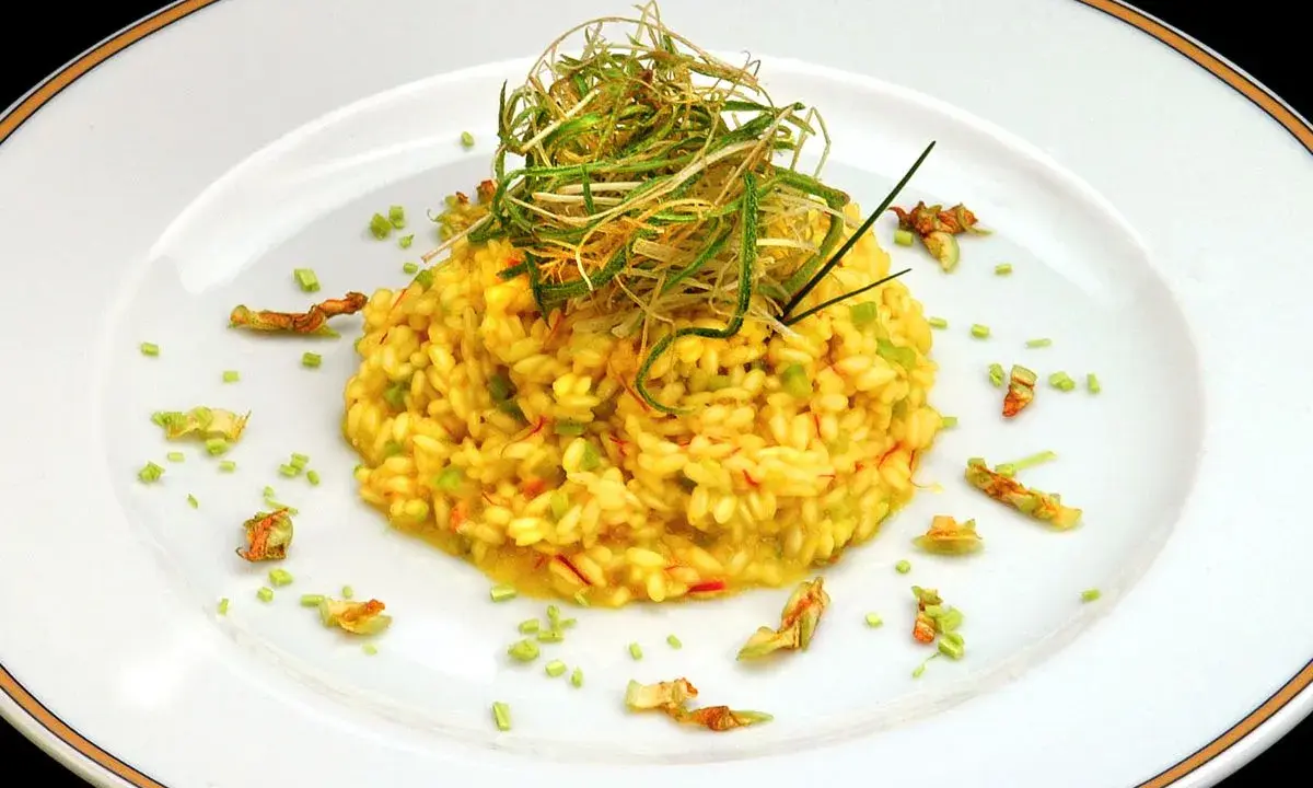 Risotto giallo intenso con fiori di zucca e zafferano, guarnito con erbe croccanti.