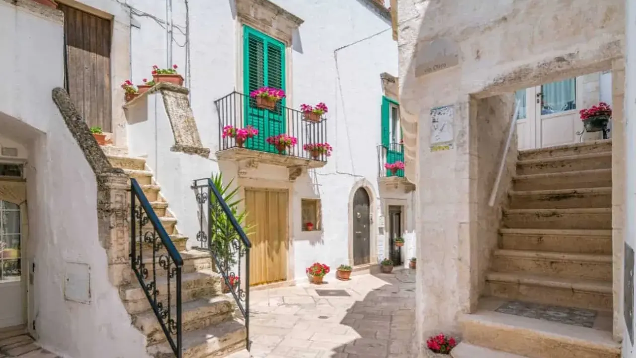 Un vicolo fiorito a Locorotondo, con scalinate in pietra bianca, balconi verdi e gerani rosa.