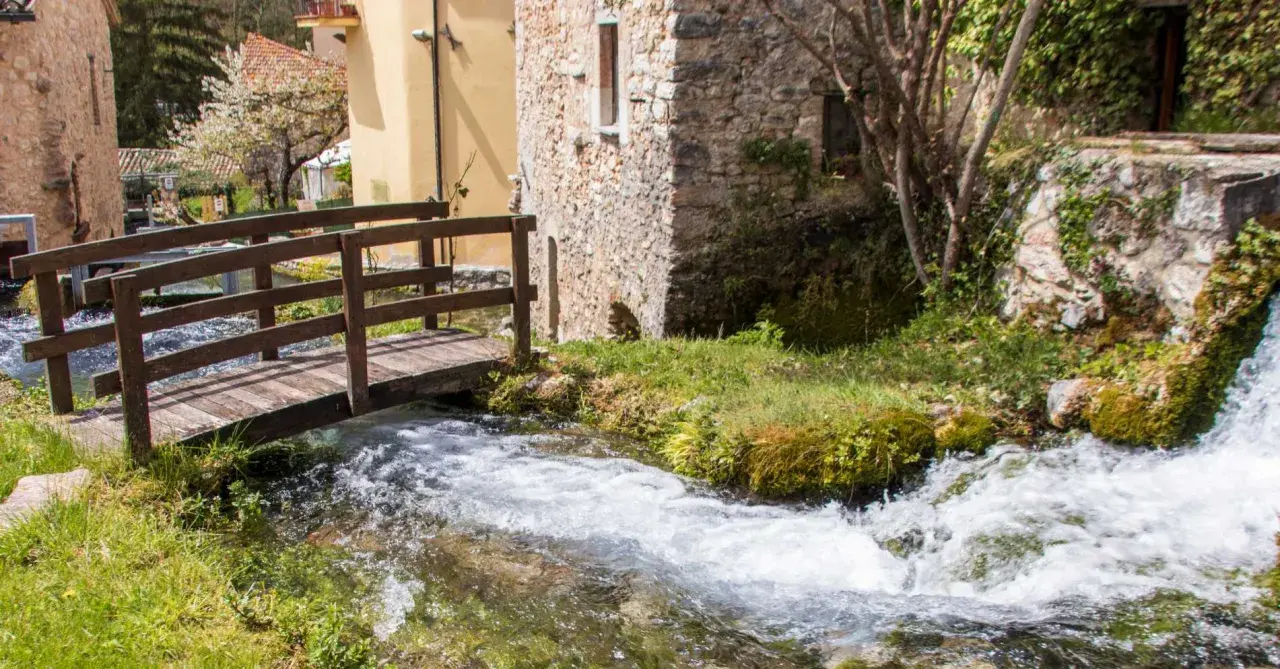 Un ponticello di legno attraversa un ruscello gorgogliante, con antiche costruzioni in pietra e un ciliegio in fiore. Un angolo di pace in Umbria dove andare.