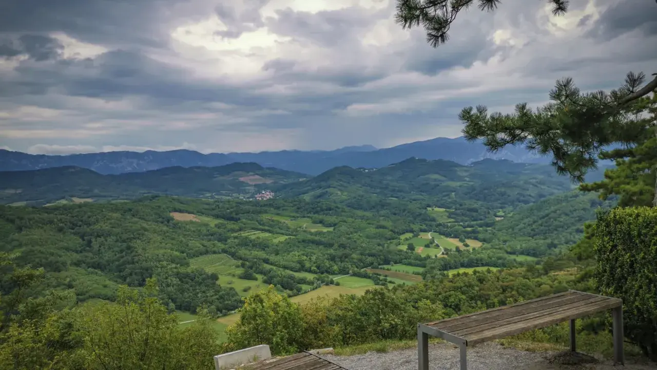 Panoramica del **Carso** con colline verdi e montagne all'orizzonte, sotto un cielo nuvoloso. Due panchine invitano alla sosta.