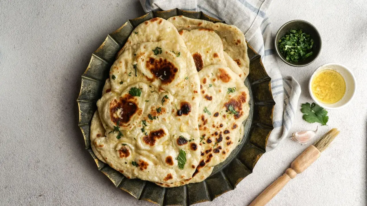 Pane in padella soffice e dorato, guarnito con erbe fresche. Accanto, ciotoline con olio e coriandolo, uno spicchio d'aglio e un pennello da cucina.