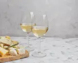 Due calici di Sauvignon Gris dorato accompagnano un tagliere di formaggi misti, tra cui erborinato e stagionato, su un tavolo grigio.