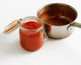 Barattolo di vetro pieno di conserva di pomodoro, accanto a una pentola con residui rossi.