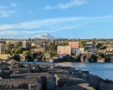 Vista sul mare con blocchi di cemento in primo piano e un panorama di case con l'Etna innevato sullo sfondo, perfetto per un itinerario a Catania.