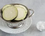 Fette di melanzane salate in un colino, pronte per spurgare. Accanto, una ciotolina di sale grosso.