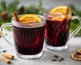 Due tazze fumanti di vin brulé, decorate con arancia e cannella, su un tavolo con spezie e rami di pino.
