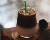 Un bicchiere di **caffè freddo** stratificato con ghiaccio e una cannuccia verde, su un tavolino di legno.