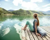 Ragazza con zaino seduta su un pontile di legno, ammira il **lago di Caldaro** e le montagne verdi sotto un cielo azzurro con nuvole bianche.