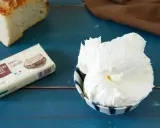 Burro montato soffice in una ciotola, accanto a una confezione di burro salato e una fetta di pane su un tavolo blu.