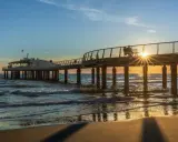 Un molo moderno si estende sul mare al tramonto, con la costa toscana mappa sullo sfondo. Le onde lambiscono la spiaggia dorata.