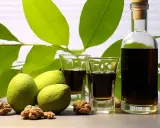 Nocino ricetta: noci verdi, liquore scuro in bicchierini e bottiglia, foglie verdi sullo sfondo.