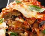 Una fetta di parmigiana di melanzane non fritte, con strati di melanzane, sugo e mozzarella filante, guarnita con basilico fresco.