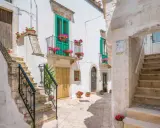 Un vicolo fiorito a Locorotondo, con scalinate in pietra bianca, balconi verdi e gerani rosa.