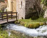 Un ponticello di legno attraversa un ruscello gorgogliante, con antiche costruzioni in pietra e un ciliegio in fiore. Un angolo di pace in Umbria dove andare.