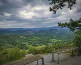 Panoramica del **Carso** con colline verdi e montagne all'orizzonte, sotto un cielo nuvoloso. Due panchine invitano alla sosta.