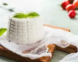 Ricotta fatta in casa, fresca e profumata, guarnita con basilico su un tagliere di legno. Sullo sfondo, pomodorini ciliegino.