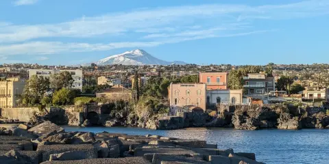 Vista sul mare con blocchi di cemento in primo piano e un panorama di case con l'Etna innevato sullo sfondo, perfetto per un itinerario a Catania.