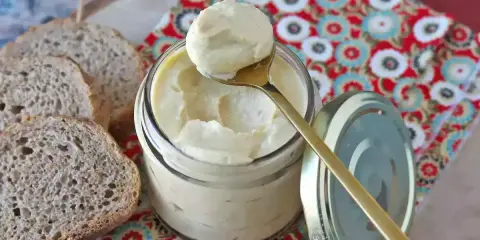 Un cucchiaio dorato preleva una generosa porzione di maionese con uovo sodo da un barattolo di vetro. Fette di pane rustico completano la scena.