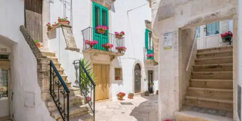 Un vicolo fiorito a Locorotondo, con scalinate in pietra bianca, balconi verdi e gerani rosa.