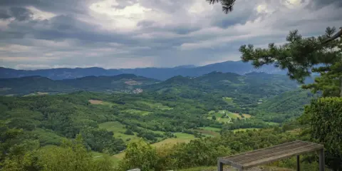 Panoramica del **Carso** con colline verdi e montagne all'orizzonte, sotto un cielo nuvoloso. Due panchine invitano alla sosta.