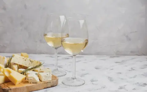Due calici di Sauvignon Gris dorato accompagnano un tagliere di formaggi misti, tra cui erborinato e stagionato, su un tavolo grigio.