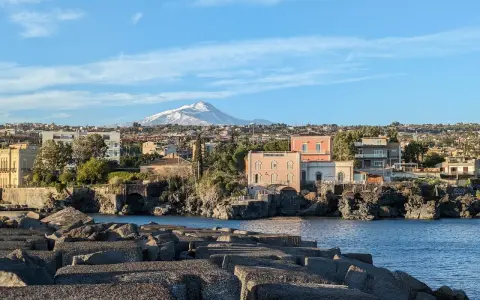 Vista sul mare con blocchi di cemento in primo piano e un panorama di case con l'Etna innevato sullo sfondo, perfetto per un itinerario a Catania.