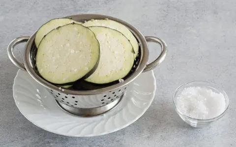 Fette di melanzane salate in un colino, pronte per spurgare. Accanto, una ciotolina di sale grosso.
