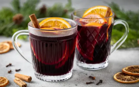 Due tazze fumanti di vin brulé, decorate con arancia e cannella, su un tavolo con spezie e rami di pino.