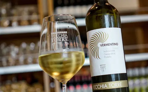 Un bicchiere di vino bianco Vermentino, frizzante e dorato, accanto alla sua bottiglia Vecchia Torre.