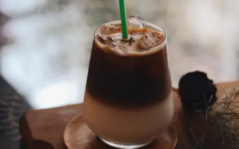 Un bicchiere di **caffè freddo** stratificato con ghiaccio e una cannuccia verde, su un tavolino di legno.