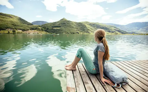 Ragazza con zaino seduta su un pontile di legno, ammira il **lago di Caldaro** e le montagne verdi sotto un cielo azzurro con nuvole bianche.