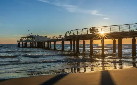 Un molo moderno si estende sul mare al tramonto, con la costa toscana mappa sullo sfondo. Le onde lambiscono la spiaggia dorata.