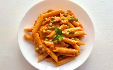Penne ai 7 peccati: un piatto di pasta cremosa con piselli e un tocco di pomodoro, guarnito con prezzemolo fresco.