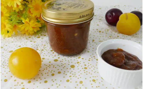 Vasetto di marmellata di susine fatto in casa, con prugne rosse e gialle e fiori gialli sullo sfondo.