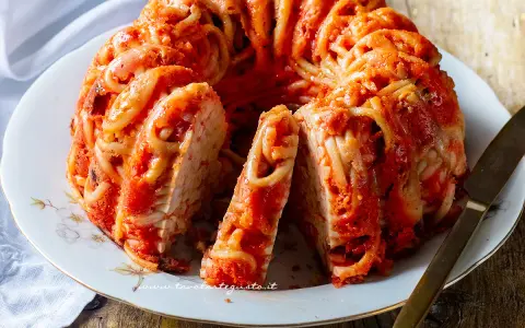 Un invitante timballo di pasta, con spaghetti avvolti in un ricco sugo rosso, servito su un piatto bianco con un coltello dorato.