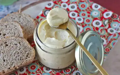 Un cucchiaio dorato preleva una generosa porzione di maionese con uovo sodo da un barattolo di vetro. Fette di pane rustico completano la scena.