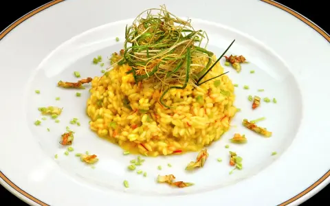 Risotto giallo intenso con fiori di zucca e zafferano, guarnito con erbe croccanti.