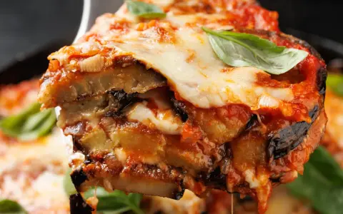 Una fetta di parmigiana di melanzane non fritte, con strati di melanzane, sugo e mozzarella filante, guarnita con basilico fresco.