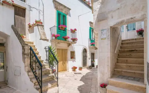 Un vicolo fiorito a Locorotondo, con scalinate in pietra bianca, balconi verdi e gerani rosa.
